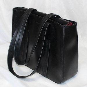 May include: A black leather tote bag with two long handles and a rectangular shape. The bag has a structured design with visible stitching and a pocket on the front. The interior appears to be lined with a red fabric.
