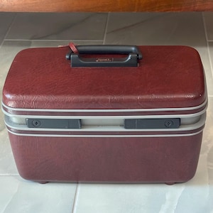 May include: A rectangular, burgundy hardshell travel vanity case with a textured finish. It features a black plastic top handle, a silver-toned trim, and two black latch closures on the front. The brand name Samsonite is visible on the handle.