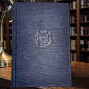 May include: A dark blue hardcover book with a textured fabric cover. Centered on the front cover is a circular embossed emblem featuring two figures standing beside a pedestal with the Latin text MENS ET MANUS inscribed around the design.