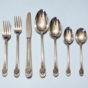 May include: A set of seven silver-toned metal utensils including two forks, one knife, and four spoons of varying sizes. Each piece features a polished finish and a decorative floral relief pattern at the base of the handle with vertical fluting.