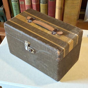 May include: A vintage rectangular travel case with a brown and tan striped pattern. The case has a brown leather handle and metal clasp. The case is sitting on a white surface, with a collection of books in the background.
