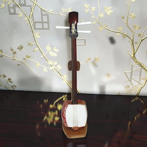 May include: A shamisen, a traditional three-stringed Japanese musical instrument, features a long, slender dark wood neck, a square body with a white skin head, and three white tuning pegs. The instrument is displayed upright on a wooden stand.