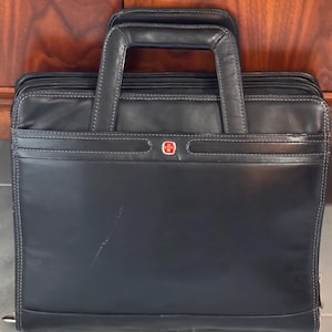May include: Black leather briefcase with a top handle and a red and white emblem. The briefcase has white stitching and a zipper closure. The bag is designed for carrying documents and other business essentials.