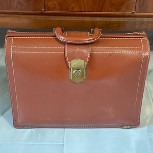 May include: A structured, rectangular brown leather briefcase featuring a top handle, white contrast stitching, and a central brass-toned metal locking mechanism with a keyhole. The bag has reinforced corners and a smooth, polished finish.