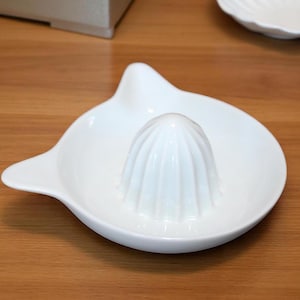 May include: A white ceramic citrus juicer with a built-in reamer and a pouring spout. The juicer is on a wooden surface. The reamer is cone-shaped with vertical ridges.