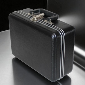 May include: A black briefcase with a textured surface, silver trim, and a black handle. The briefcase has a metal clasp and hinges. The case is rectangular and appears to be made of a durable material.