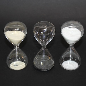 May include: Three clear glass hourglasses arranged in a row. The left hourglass contains beige sand, the center hourglass contains black sand, and the right hourglass contains white sand. Each hourglass has a rounded top and bottom bulb.