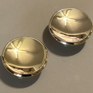 May include: Two shiny, gold-colored, round cabinet knobs. Each knob has a reflective surface and a white, star-like design on the top. The knobs are set against a gray background, casting shadows.