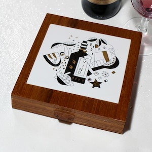 May include: Wooden box with a white tile top featuring a black and gold illustration of a wine bottle and other wine-related elements. The box has a dark brown finish and a small wooden latch.