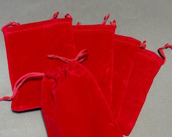 May include: A collection of red velvet drawstring bags. The bags are a vibrant shade of red, with a soft, plush texture. The drawstring closure is made of a matching red cord. The bags are arranged on a neutral gray surface.