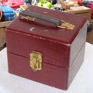 May include: A vintage burgundy box with a brown handle and gold-colored hardware. The box is rectangular with a latch closure and shows signs of wear. The handle is attached with gold-colored metal brackets.