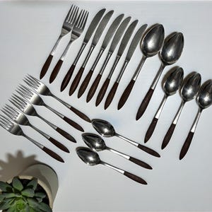 May include: A collection of vintage silverware, including forks, knives, and spoons. The utensils have stainless steel heads and dark brown wooden handles. The arrangement is on a white surface, with a small potted plant in the corner.