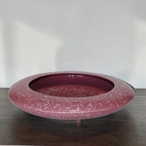 May include: A round, low-profile ceramic bowl with a wide, flat rim. The bowl features a deep mauve or dusty rose base color with a speckled, white, and light gray mottled pattern across the entire surface, supported by small, subtle feet.