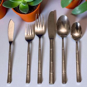 May include: A set of six vintage silverware pieces, including a butter knife, two forks, a knife, and two spoons. The silverware has a bamboo-style handle design and a silver finish. The set is arranged on a light-colored surface.