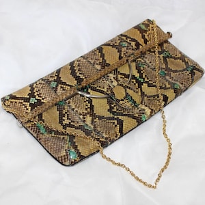 May include: A snakeskin-patterned clutch with a gold chain strap. The clutch features a mix of brown, tan, and green tones, with a silver-tone metal clasp. The chain is a delicate gold color, adding a touch of elegance to the accessory.