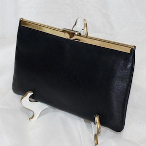 May include: A rectangular black clutch bag featuring a smooth finish and a metallic gold-toned frame. The top of the bag is secured with a gold-toned metal clasp closure that runs along the entire length of the upper edge of the accessory.