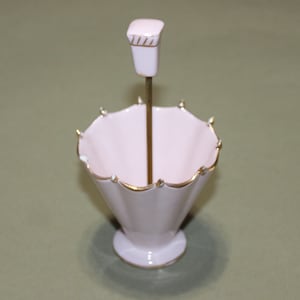 May include: A pink ceramic ring holder shaped like an umbrella with a gold trim. The holder has a small, pink ceramic knob on top of a gold metal rod.