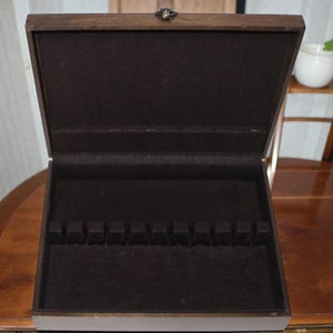 May include: Open wooden cutlery box with a dark brown felt interior. The box has a hinged lid with a metal clasp. The interior features slots for holding silverware. The box is sitting on a wooden table.