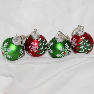 May include: Four Christmas ornaments in red and green. Each glass ball has a silver cap and a twine loop for hanging. The ornaments are decorated with white snowflakes and green Christmas trees.