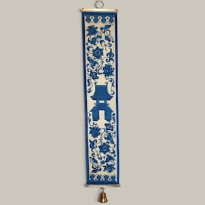 May include: A vertical fabric wall hanging featuring a blue and white floral pattern with a central pagoda illustration. The textile is suspended from a gold-toned metal rod with a circular loop at the top and a small brass bell attached at the bottom.