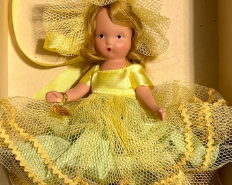 Puede incluir: Una muñeca vintage con cabello rubio y un vestido amarillo. El vestido de la muñeca está hecho de un material similar al satén y está cubierto con una red amarilla. La muñeca lleva un gran lazo amarillo y una pulsera dorada. La muñeca está en una caja.