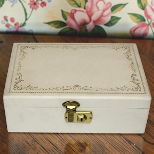 May include: A rectangular, off-white jewelry box with a gold-colored clasp and decorative trim. The box has a hinged lid and a lock. The trim features a delicate, repeating floral pattern around the edges of the lid.