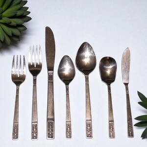 May include: A set of seven silver-toned stainless steel flatware pieces arranged in a row, including two forks, one dinner knife, two spoons, one smaller spoon, and one butter knife, all featuring a decorative embossed pattern on the handles.