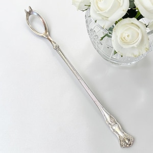 May include: A long, silver-toned metal olive spoon featuring an oval-shaped bowl with two small prongs at the tip. The handle is slender and decorated with embossed floral and vine-like patterns at the base and near the bowl attachment point.