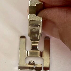 May include: A silver-colored sewing machine presser foot. The foot has a rectangular top with a curved edge, a central section with two small holes, and two flat, parallel bars extending downward. The foot is designed to attach to a sewing machine.