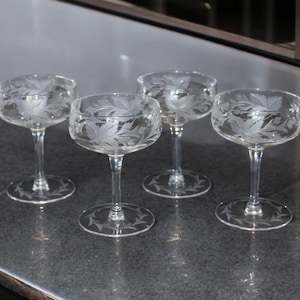 May include: Four clear champagne coupe glasses with etched leaf designs. Each glass has a round bowl, a slender stem, and a circular base with a decorative etched pattern. The glasses are arranged on a dark, reflective surface.