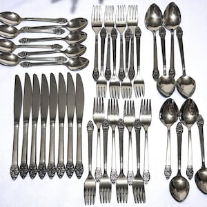 May include: A collection of silver-colored flatware, including spoons, forks, and knives, arranged on a white surface. The silverware features ornate floral detailing on the handles. The set includes multiple pieces of each utensil type, suitable for a formal dining setting.