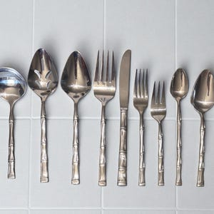 May include: A collection of stainless steel silverware with bamboo-style handles. The set includes a butter knife, soup ladle, slotted spoon, serving spoon, forks, knives, and spoons. The silverware is arranged on a light gray tiled surface.