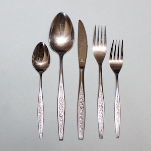 May include: A set of five silver-toned flatware pieces, including a knife, fork, and three spoons. The spoons have ornate designs.
