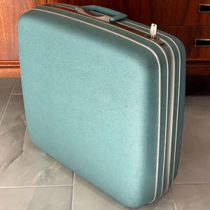 May include: A teal hardshell suitcase with a textured finish, featuring a matching teal top handle with silver-toned accents, a silver-toned metal trim along the edges, and a small silver key inserted into the locking mechanism on the top side.