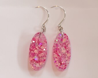 May include: A pair of dangle earrings with silver-colored hooks. Each earring features an oval-shaped design filled with pink glitter and iridescent flakes. The earrings are set against a white background.