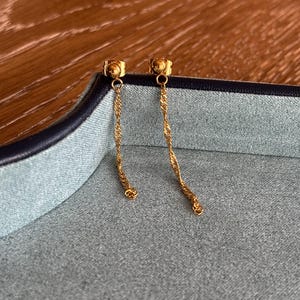 May include: A pair of gold-colored drop earrings. Each earring features a small stud with a twisted chain that hangs down. The earrings are displayed on a gray surface with a dark blue border, set against a wooden background.