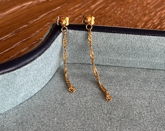 May include: A pair of gold-colored drop earrings. Each earring features a small stud with a twisted chain that hangs down. The earrings are displayed on a gray surface with a dark blue border, set against a wooden background.