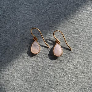May include: A pair of gold-colored drop earrings with pear-shaped, pale pink gemstones. The earrings hang from curved gold-colored hooks. The background is a textured, muted blue-gray fabric.