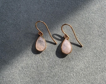 May include: A pair of gold-colored drop earrings with pear-shaped, pale pink gemstones. The earrings hang from curved gold-colored hooks. The background is a textured, muted blue-gray fabric.