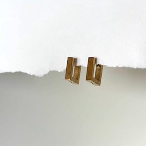 May include: A pair of gold-colored, rectangular hoop earrings. Each earring has a geometric, open design. The earrings are displayed on a white background.