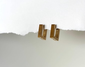 May include: A pair of gold-colored, rectangular hoop earrings. Each earring has a geometric, open design. The earrings are displayed on a white background.