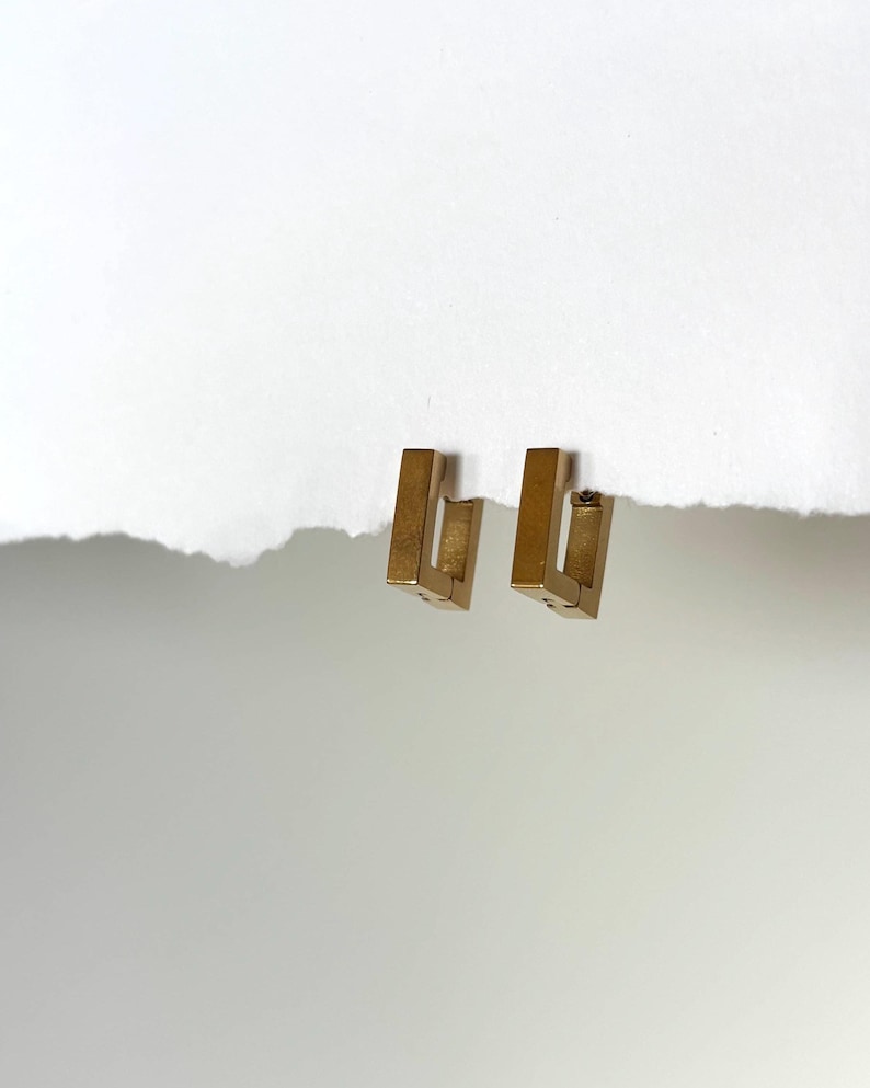May include: A pair of gold-colored, rectangular hoop earrings. Each earring has a geometric, open design. The earrings are displayed on a white background.