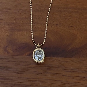 May include: A gold-toned necklace featuring a delicate ball chain and a single oval-shaped pendant. The pendant consists of a clear, faceted stone set within a smooth, polished gold-colored metal bezel frame that catches the light.