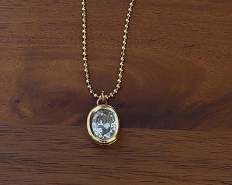 May include: A gold-toned necklace featuring a delicate ball chain and a single oval-shaped pendant. The pendant consists of a clear, faceted stone set within a smooth, polished gold-colored metal bezel frame that catches the light.