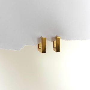 May include: A pair of gold-colored, rectangular hoop earrings. The earrings have a simple, geometric design with a vertical bar and a small, square clasp. The earrings are displayed against a white background.