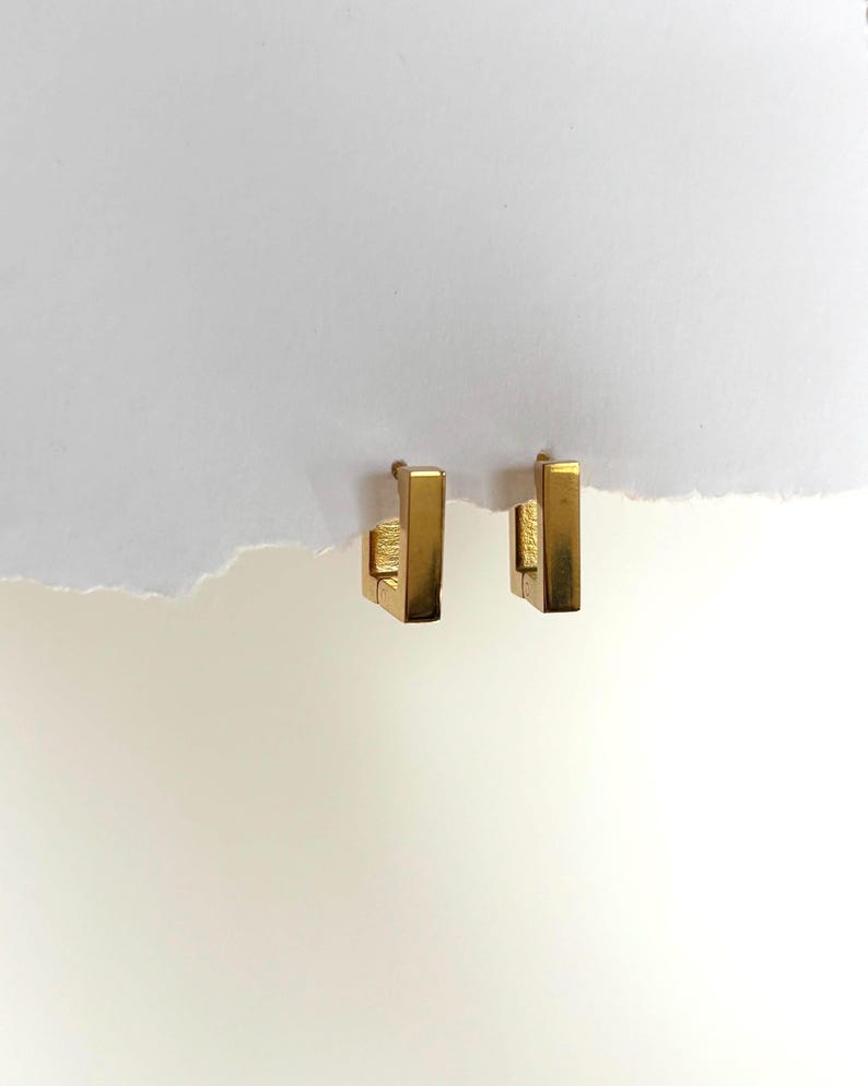 May include: A pair of gold-colored, rectangular hoop earrings. The earrings have a simple, geometric design with a vertical bar and a small, square clasp. The earrings are displayed against a white background.