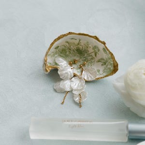 May include: An oyster shell dish with a gold rim and green floral design holds white petal earrings with gold accents. A bottle of Dolce & Gabbana Light Blue perfume is in the foreground.