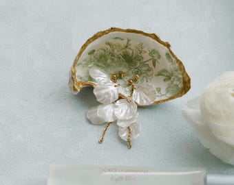 May include: An oyster shell dish with a gold rim and green floral design holds white petal earrings with gold accents. A bottle of Dolce & Gabbana Light Blue perfume is in the foreground.