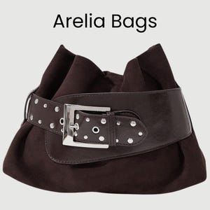 May include: A dark brown suede handbag with a wide, studded belt and a silver buckle. The bag has a gathered design and the text "Arelia Bags" is displayed at the top.