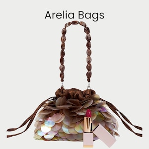 Puede incluir: Un bolso con asa de cuentas marrones y lentejuelas iridiscentes, con cierre de cordón. Se combina con un labial rosa con tubo dorado y tapa rosa a juego. El texto "Arelia Bags" está en la parte superior.